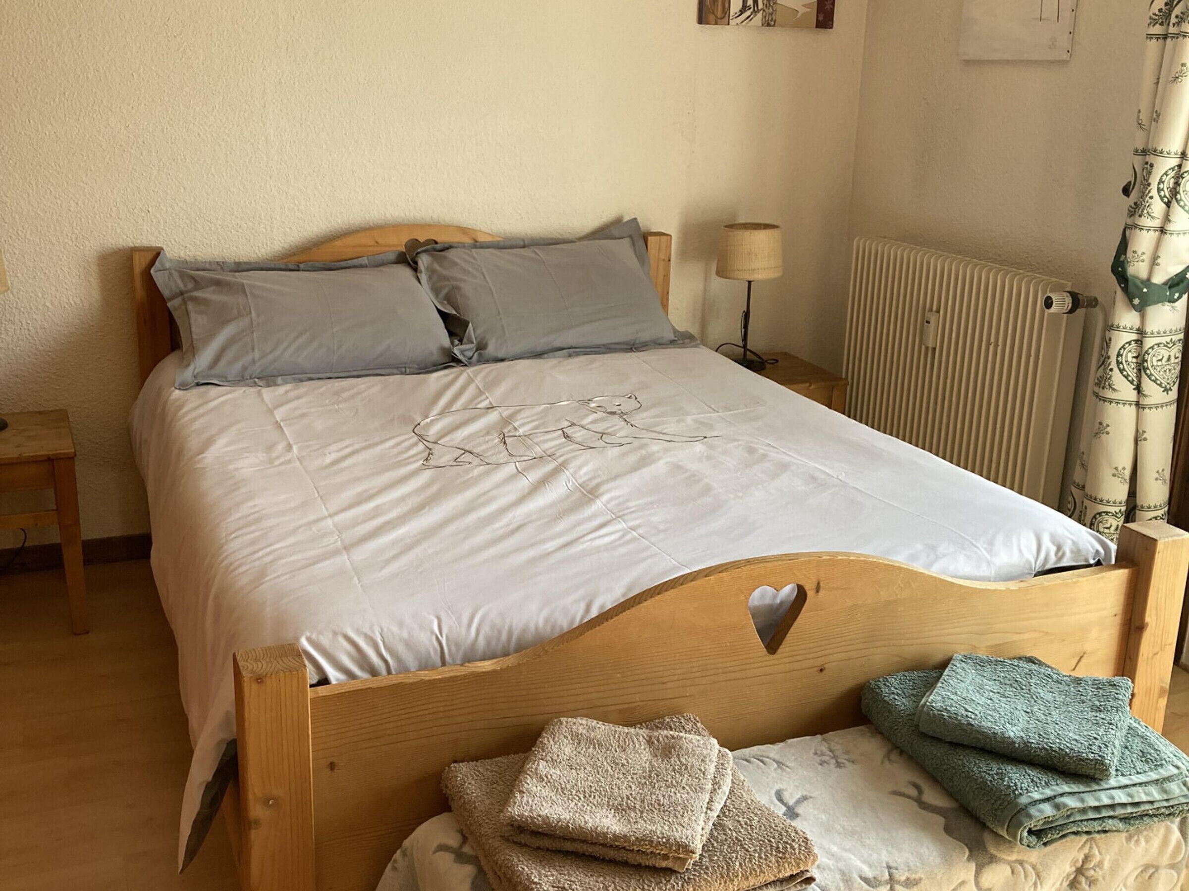 A bedroom with a wooden headboard, a bed with grey and white bedding, a bedside lamp, a radiator, and a window with curtains.