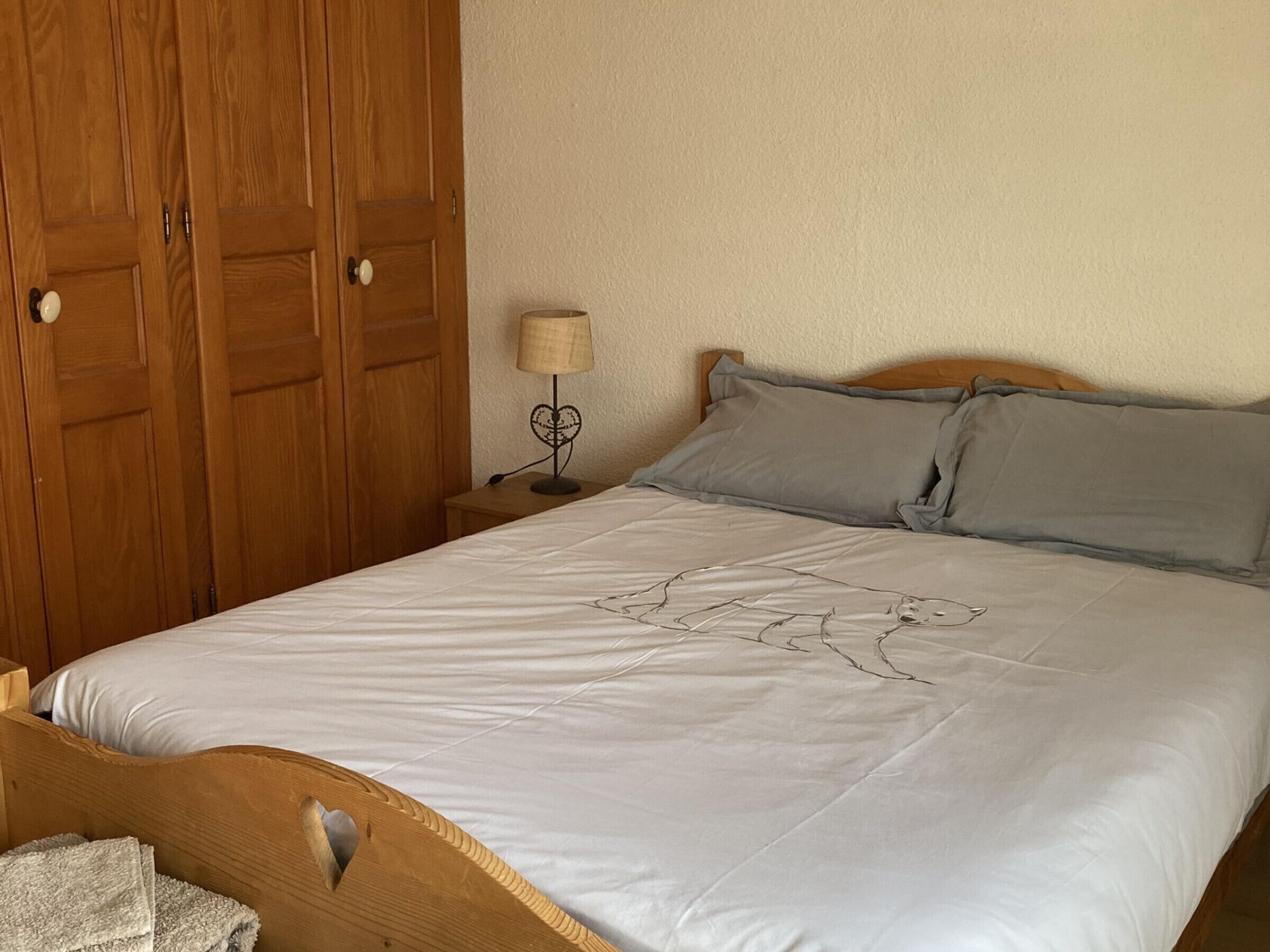 A bedroom with a wooden bed frame, a white mattress, grey pillows, a bedside table with a lamp, and a wooden wardrobe.