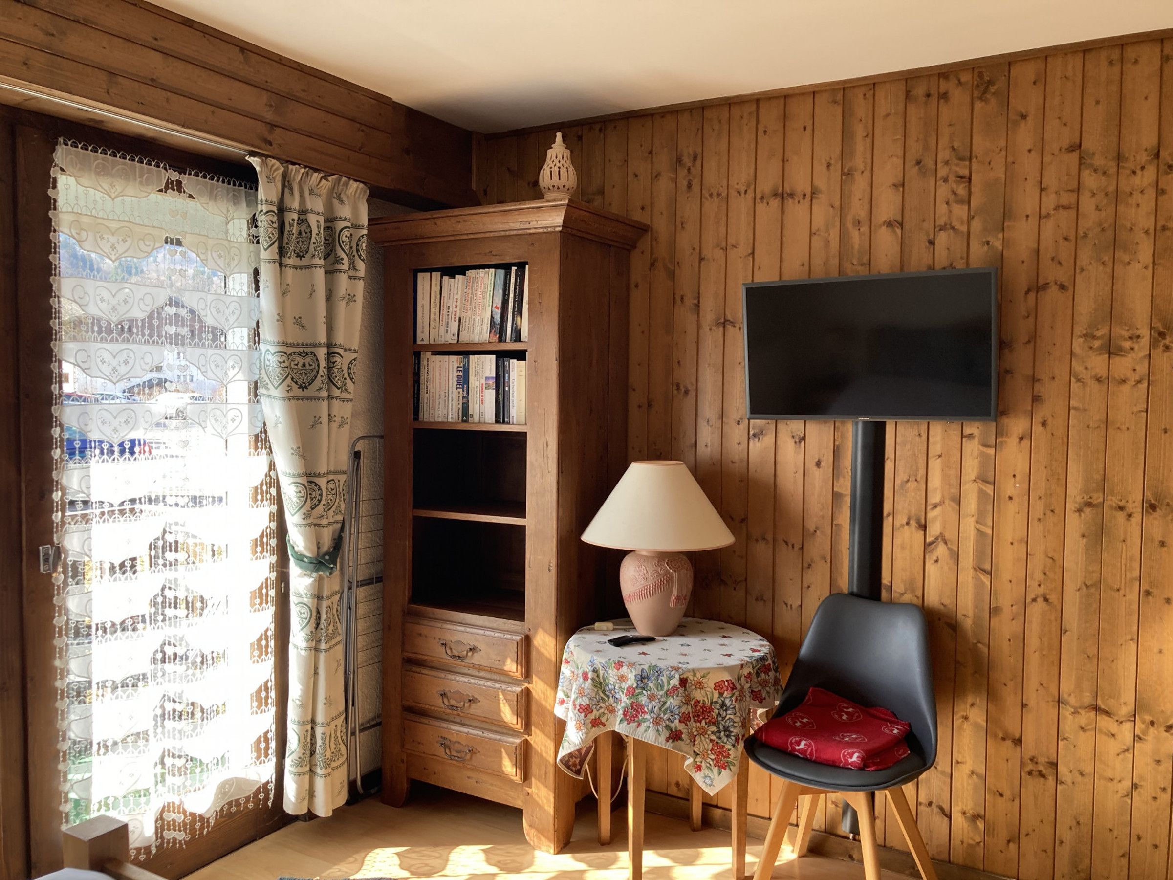 Ein Zimmer mit Holzvertäfelung, einem Bücherregal, einem Tisch mit Lampe, einem Sessel und einem an der Wand befestigten Fernseher.
