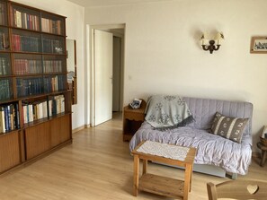 A living room with a wooden bookshelf, a sofa with a blanket, a small wooden table, and a wall-mounted lamp. - Cozy apartment for 2 with Wifi in Samoëns (Samoëns)