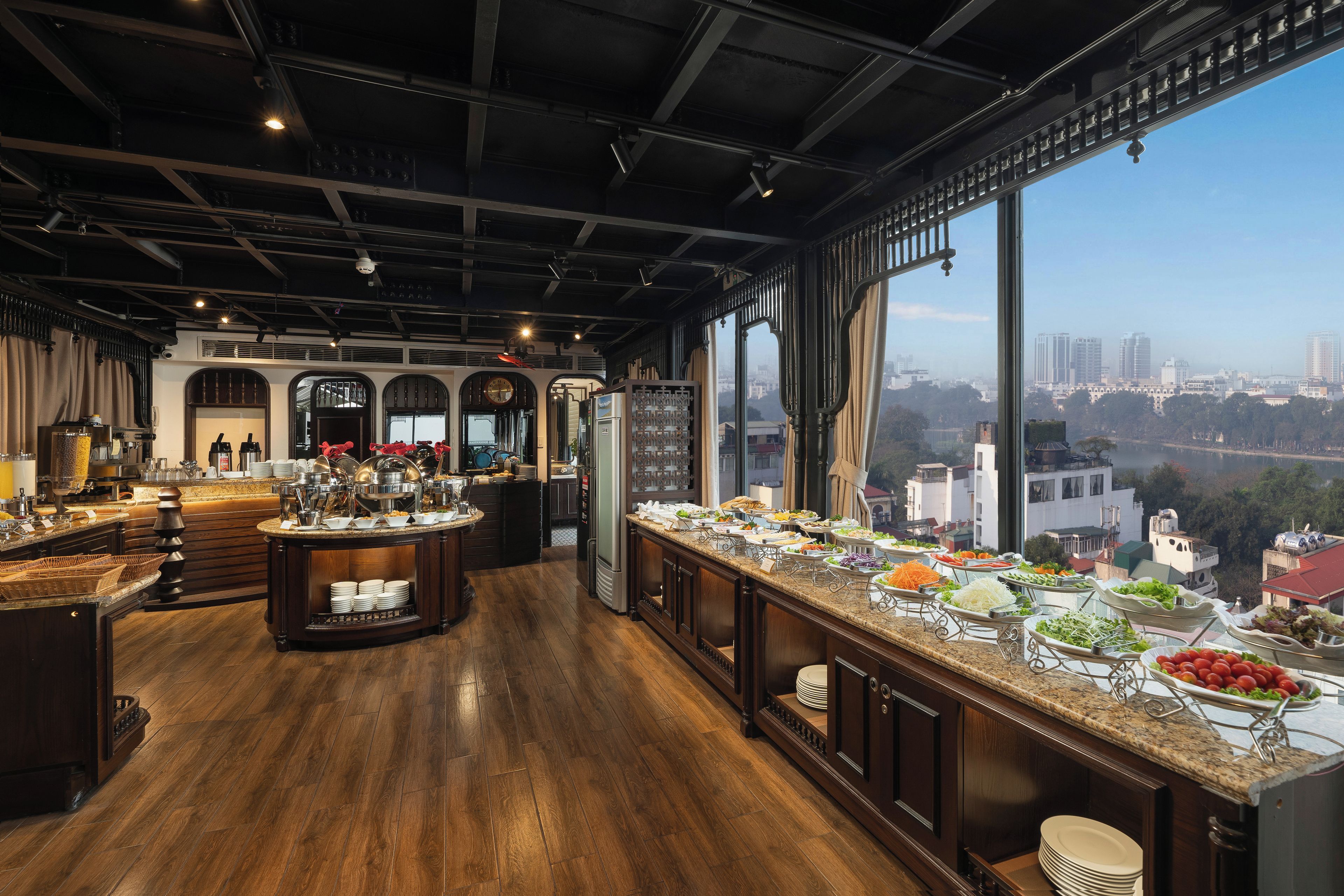 A spacious kitchen with a buffet setup, large windows offering a city view, and a modern industrial ceiling design.