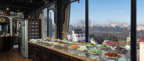 A buffet setup with a variety of food items, a clock on the wall, and a view of a cityscape through large windows.