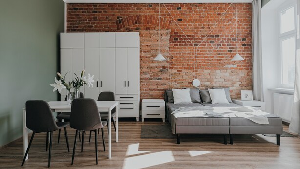 Une chambre moderne avec un mur en briques, un lit, des tables de chevet, une armoire et un coin repas avec des chaises et une table.