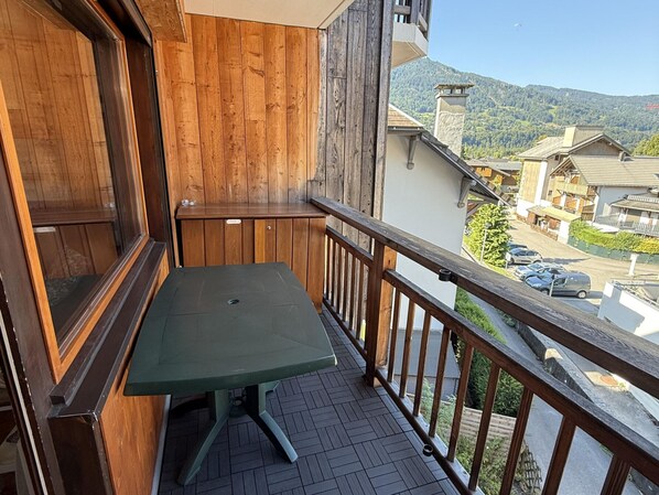 Un balcon avec une balustrade en bois, une table verte et une vue sur une rue résidentielle et des montagnes.