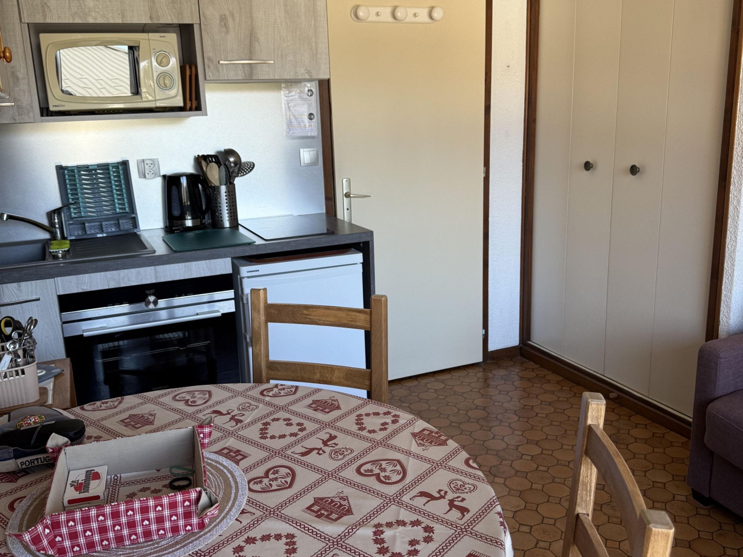 A kitchen with a microwave, coffee maker, and a refrigerator. There is a dining table with a patterned tablecloth and a chair.