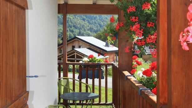 Puerta de madera que da a un balcón con mesa y sillas, rodeado de flores rojas.