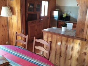 A wooden cabin interior with a kitchen area, a dining table with chairs, and a lamp.