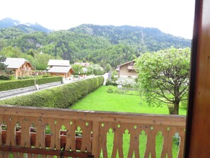 Vue depuis un balcon doté d’une balustrade en bois, donnant sur un jardin bien entretenu, avec des haies soigneusement taillées et une variété d’arbres. Au loin, on aperçoit des bâtiments à l’architecture traditionnelle, et une chaîne de montagnes se profile à l’horizon.