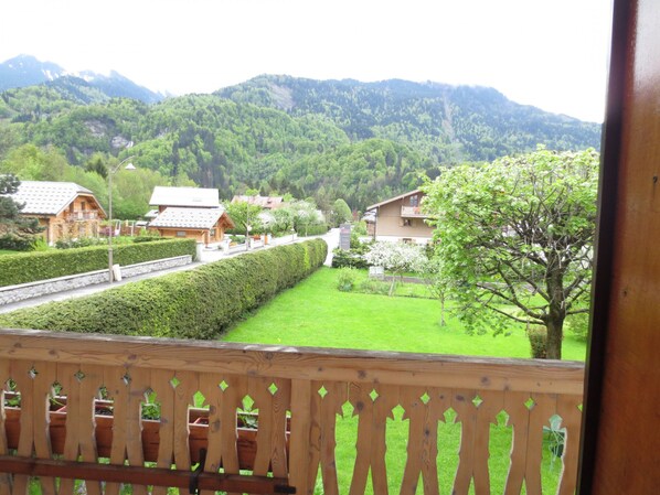 A view from a balcony with a wooden railing, overlooking a well-maintained garden with neatly trimmed hedges and a variety of trees. In the background, there are buildings with traditional architecture, and a mountain range is visible.