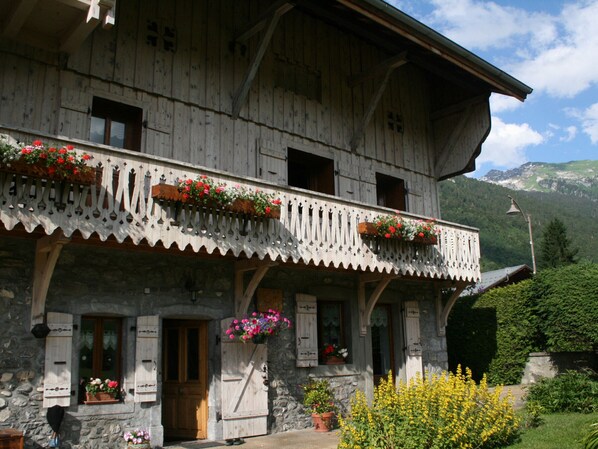 A traditional wooden chalet with balconies adorned with flower boxes, stone walls, and a mountainous backdrop.