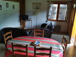 A dining area with a striped tablecloth, wooden chairs, a television on a stand, and a window with curtains. - Cozy House in Samoëns - 2 Bedrooms, Wi-Fi, Sleeps 6 (Samoëns)