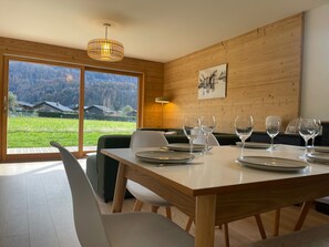 A dining area with a wooden table, chairs, and a view of a green landscape through large windows. - Spacious apartment in Samoëns - 8 people with Wifi (Samoëns)