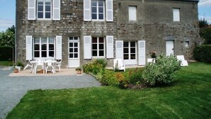 A stone building with multiple windows and shutters, a paved patio with outdoor furniture, and a well-maintained lawn.