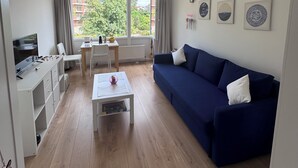 A modern living room with a blue sofa, a white coffee table, and a dining area with a table and chairs. There is a TV on a stand and a window with curtains. - Bright Blueberry Studio (Bruxelles)