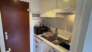 A compact kitchen with white cabinets, a built-in oven, a sink, and a dish rack. There is a small dishwasher and a door with a handle. - Bright Blueberry Studio (Bruxelles)