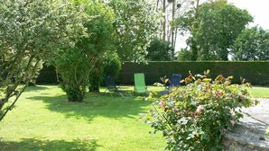 Un jardin bien entretenu, avec une pelouse, une haie et un mur de pierres. On y trouve deux chaises de jardin vertes et une petite table.