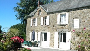 Un bâtiment en pierre avec une allée gravillonnée, une terrasse avec une table et des chaises, et des plantes en fleurs.