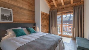 Un dormitorio con techo de madera, cama con ropa de cama blanca y verde azulado, un banco y vistas a una terraza de madera exterior.