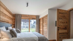 Un dormitorio moderno con una cama grande, paneles de madera en las paredes, una puerta corrediza de vidrio a un balcón y una mesita de noche blanca.