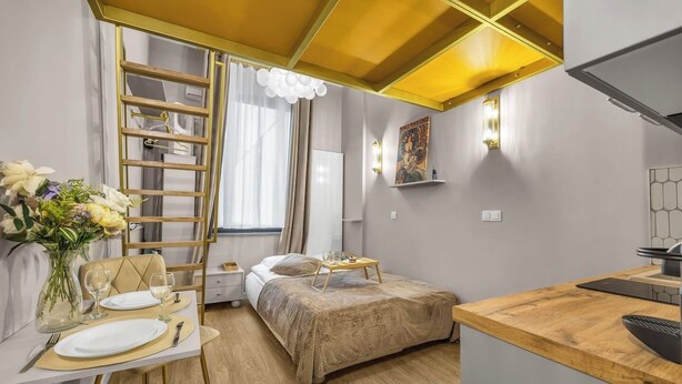 A compact hotel room with a bed, a small desk, a chair, a dining table set for two, a ladder, and a wall-mounted shelf with a decorative item. - Warsaw Praga Studio – Central Access & Parking (Warszawa)