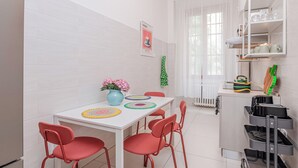 Une cuisine compacte aux carreaux blancs, une table à manger avec des chaises rouges, un meuble blanc avec des étagères et une fenêtre ornée de rideaux blancs.