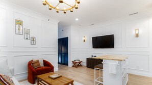 Un salon moderne comprenant un canapé, un fauteuil rouge, une table basse en bois, un téléviseur fixé au mur et un coin cuisine avec un comptoir et des placards.