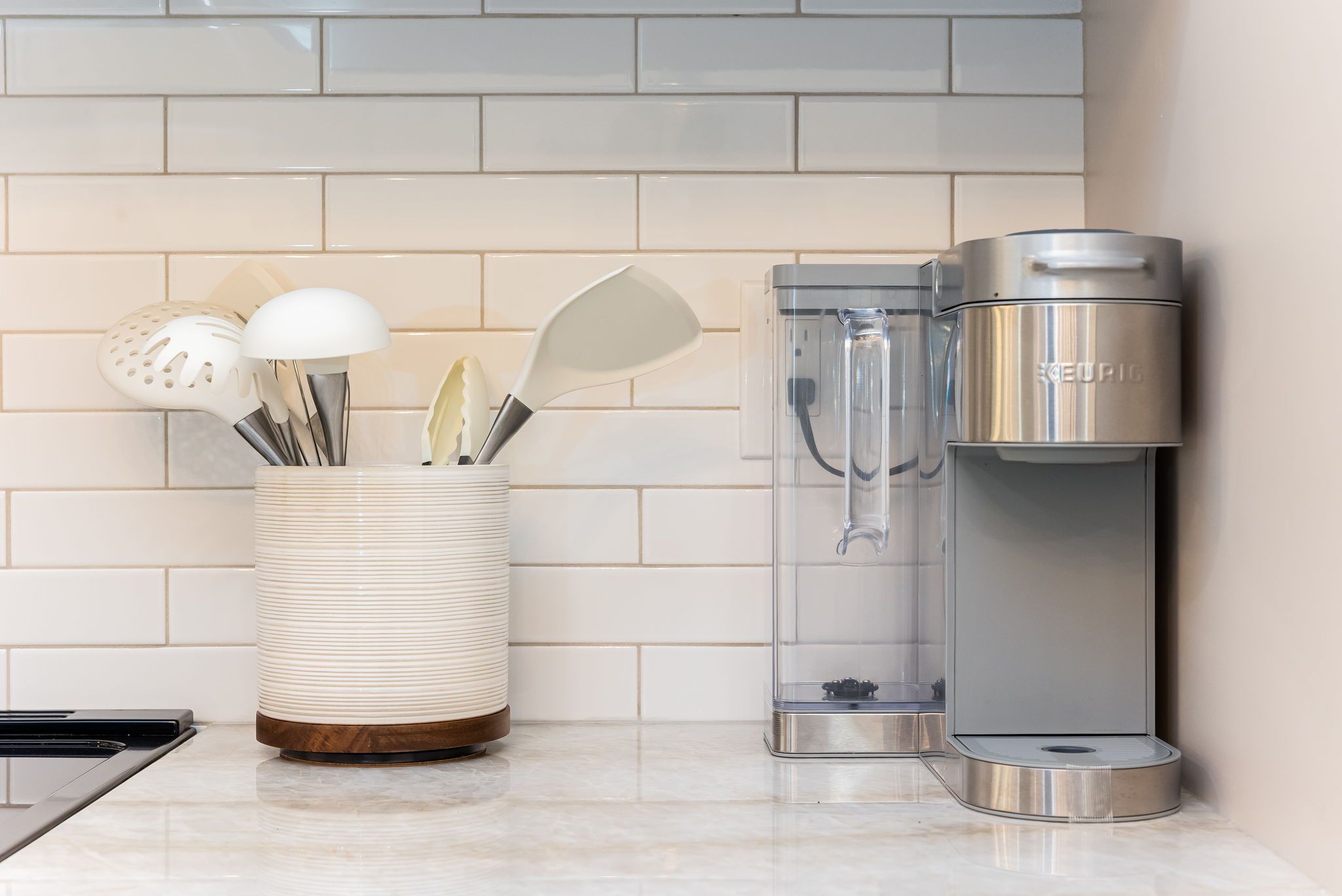 A kitchen with a white tiled backsplash, a stainless steel coffee maker, and a utensil holder with spatulas and a slotted spoon.