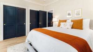Une chambre avec un grand lit, du linge de lit blanc, un plaid orange et deux tableaux encadrés accrochés au mur.