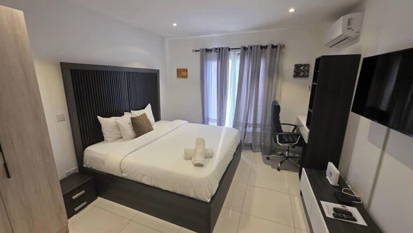 A modern hotel room with a large bed, a desk with a chair, a flat-screen TV, and a window with curtains. - Cozy Nuggets at Lennox (Accra)