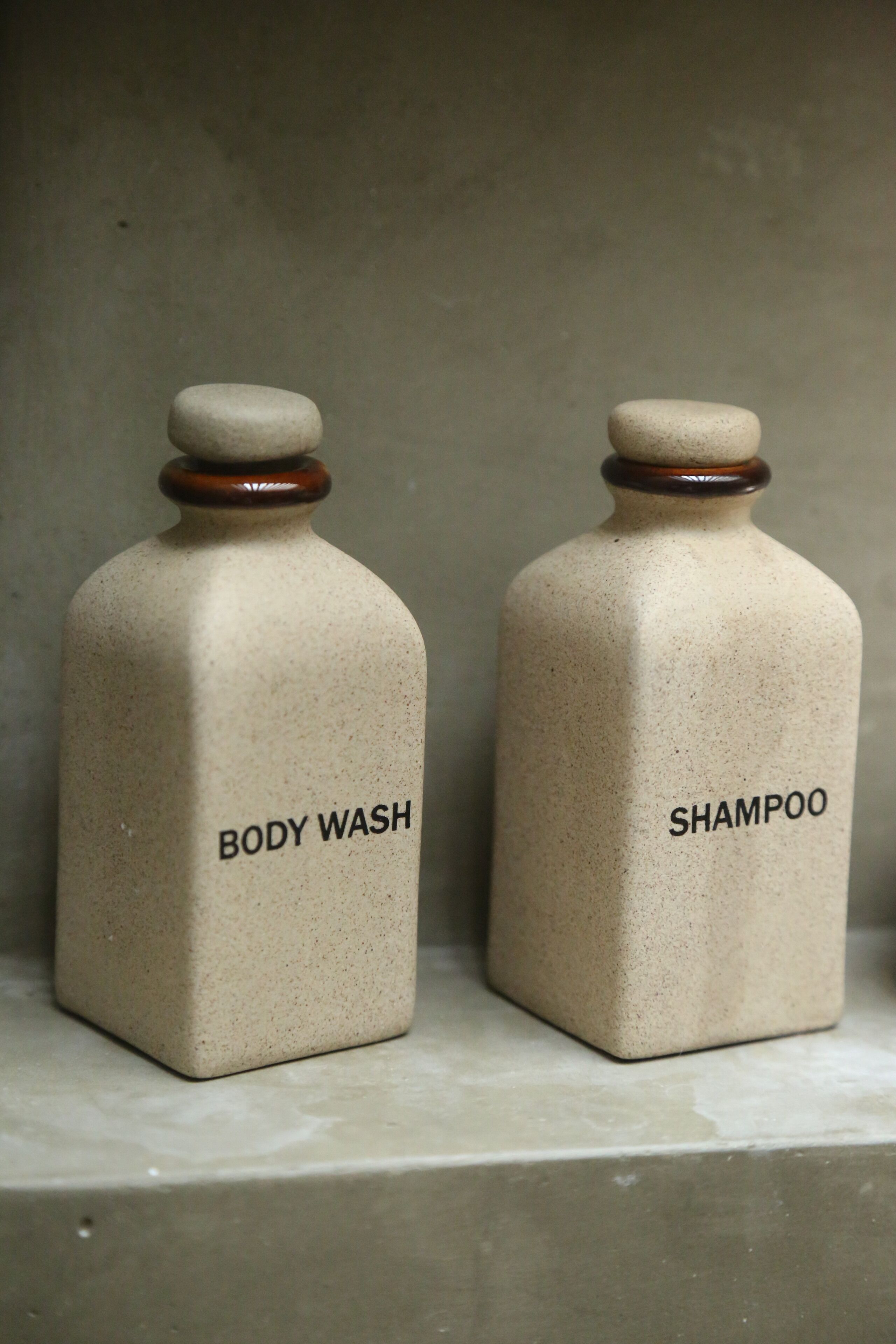 Two ceramic bottles labeled "BODY WASH" and "SHAMPOO" on a shelf.