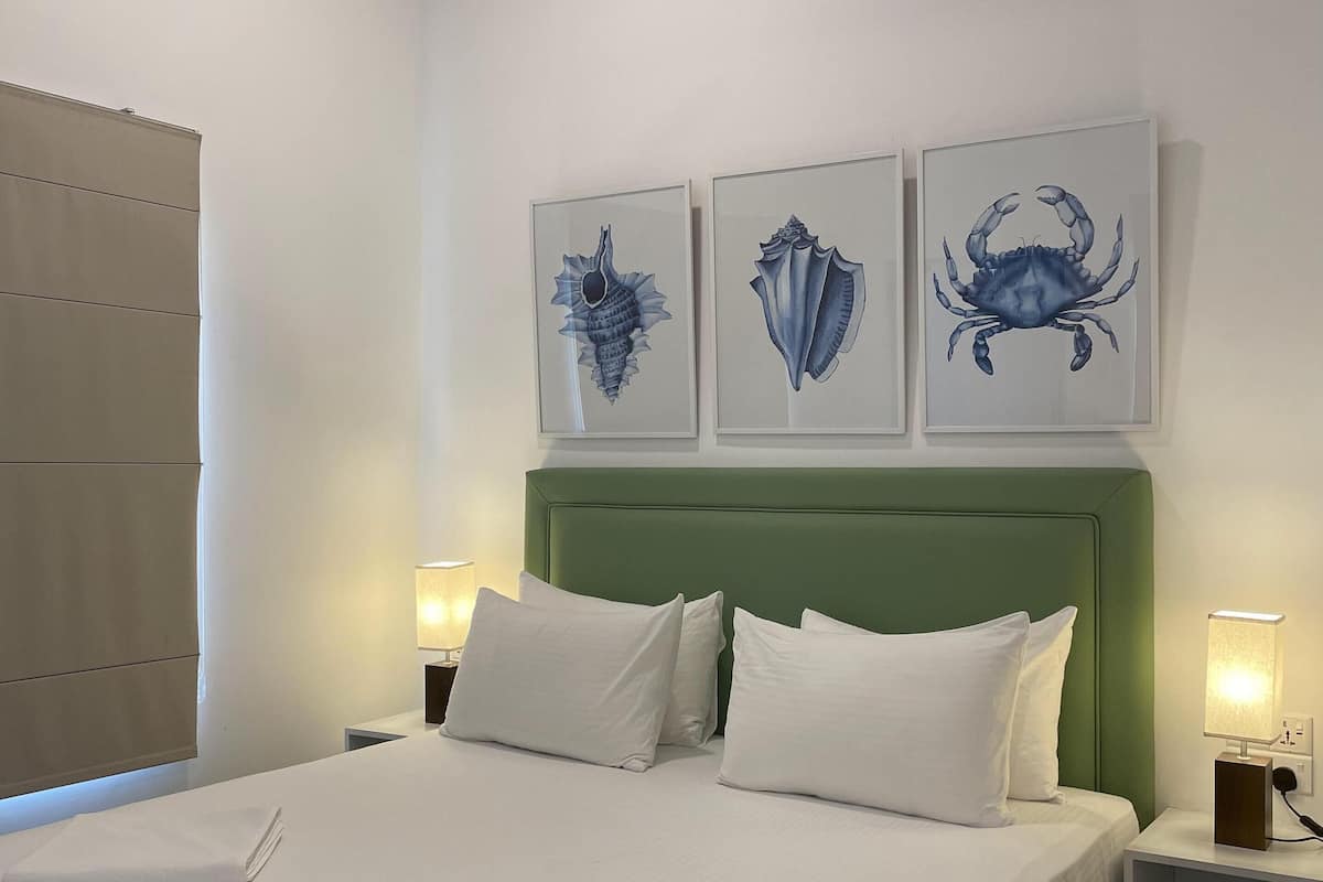 A neatly made bed with white linens and pillows, a green upholstered headboard, and three framed artworks above it.