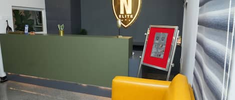 A reception area with a green counter, a yellow sofa, a wall-mounted sign with 'N ELITE', and a red information stand.
