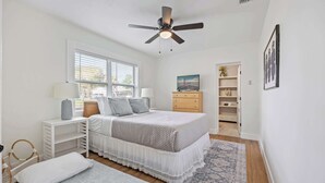 Un dormitorio con cama, mesitas de noche, ventilador de techo, ventana, cómoda y puerta que da a otra habitación.