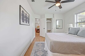 A bedroom with a bed, a ceiling fan, a window with blinds, a door leading to a bathroom, and a framed picture on the wall.