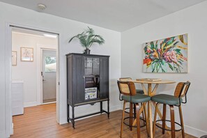 A dining area with a round wooden table and two chairs, a cabinet with a plant, and a colorful floral painting on the wall.