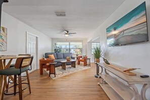 A spacious living room with a sofa, coffee table, and chairs, a dining area with a table and chairs, and a large painting of a bridge on the wall.