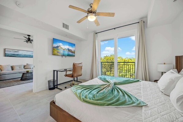 A hotel room with a bed, a desk, a chair, a ceiling fan, and a large window with a balcony view.
