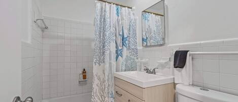 A bathroom with a bathtub, a shower curtain with a marine design, a sink with a mirror above it, and a toilet.