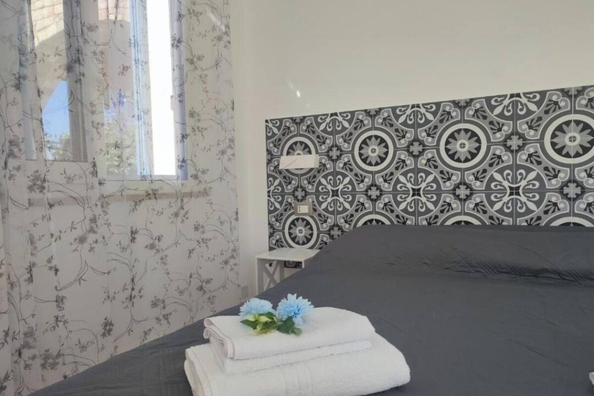 A bedroom with a patterned headboard, a window with floral curtains, and a neatly made bed with a towel and flowers.