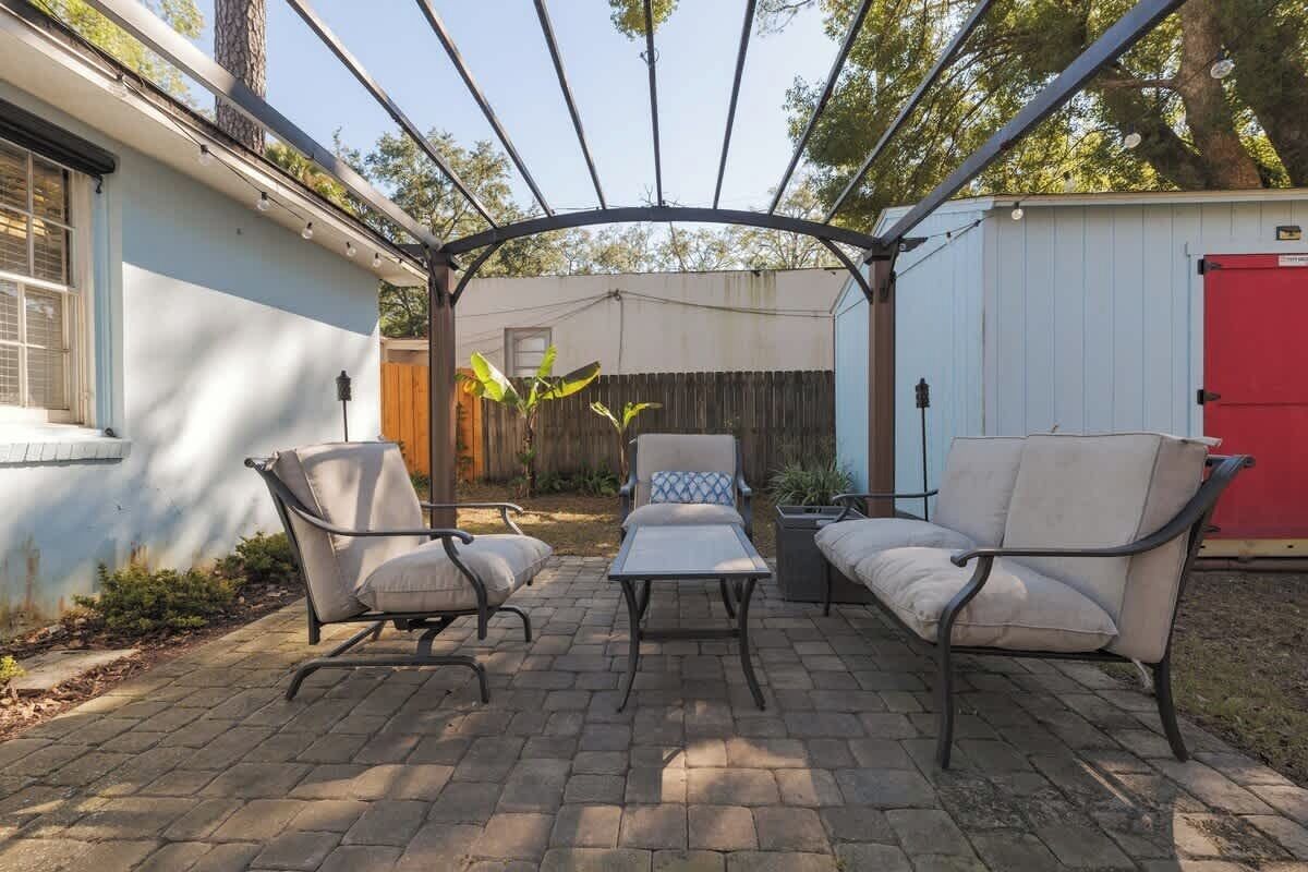 Un patio con una pérgola, dos sillas acolchadas, una mesita y un cobertizo rojo al fondo.