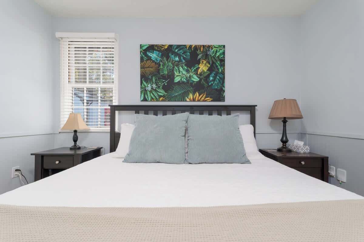 A bedroom with a bed, two bedside tables, a window with blinds, and a wall-mounted artwork.