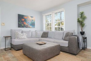 Un salon avec un canapé d’angle gris, une table basse, un tableau accroché au mur, une fenêtre avec des stores et une plante en pot.