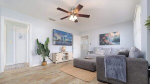 Un salon moderne avec un canapé gris, un ventilateur de plafond, une grande plante en pot et deux tableaux accrochés au mur.