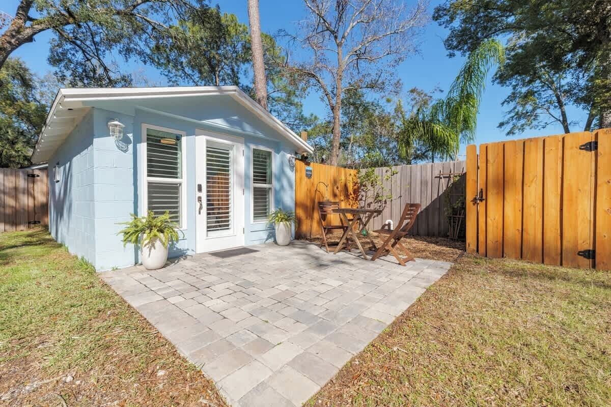 A small, light blue house with a patio, a wooden fence, and a tree in the background.