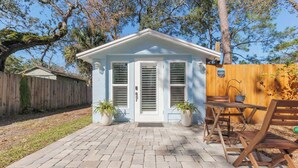 Une petite maison bleu clair avec une porte d’entrée, deux fenêtres et une terrasse en briques.