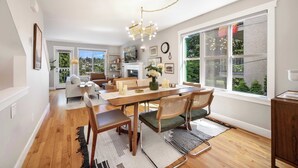 Uma sala de jantar com mesa de madeira, cadeiras e um candelabro. Há quadros nas paredes e uma vista para o verde lá fora.