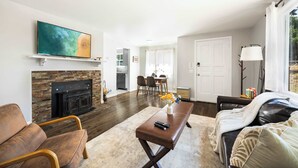 A living room with a fireplace, a TV mounted on the wall, a dining area with chairs, and a sofa with a blanket. - Vintage Charm 3BR Home w,Yard, Pets Welcome (Shoreline)