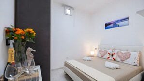 Hotellihuone, jossa on sänky, yöpöytä pullolla ja laseilla, maljakko kukkineen, meritähtikoriste ja taulu seinällä.