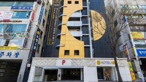 Ein Hotel mit auffälliger gelb-schwarzer Fassade, einem gut sichtbaren 'P'-Schild für die Parkplätze und verschiedenen Geschäften im Erdgeschoss.