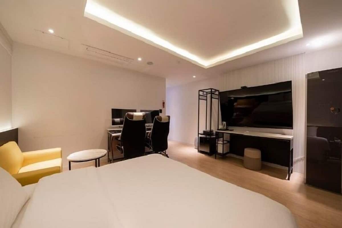 A modern hotel room with a large bed, a desk with two chairs, a TV, and a seating area with a yellow chair.